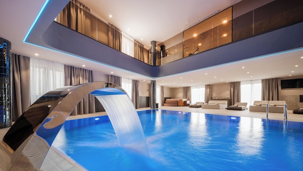 Una piscina interior moderna con caída de agua, un área de descanso con asientos y un techo alto con iluminación empotrada.