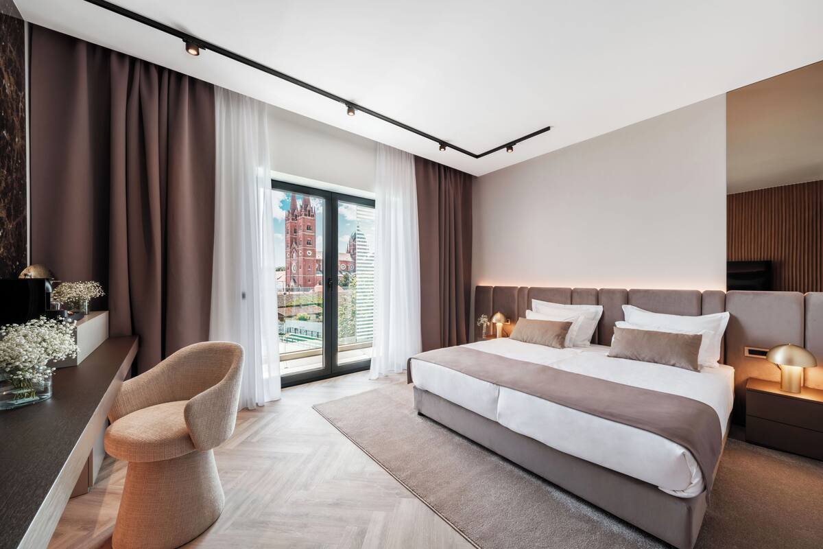 A modern bedroom with a large bed, a chair, a nightstand, and a view of a cityscape through a glass door.