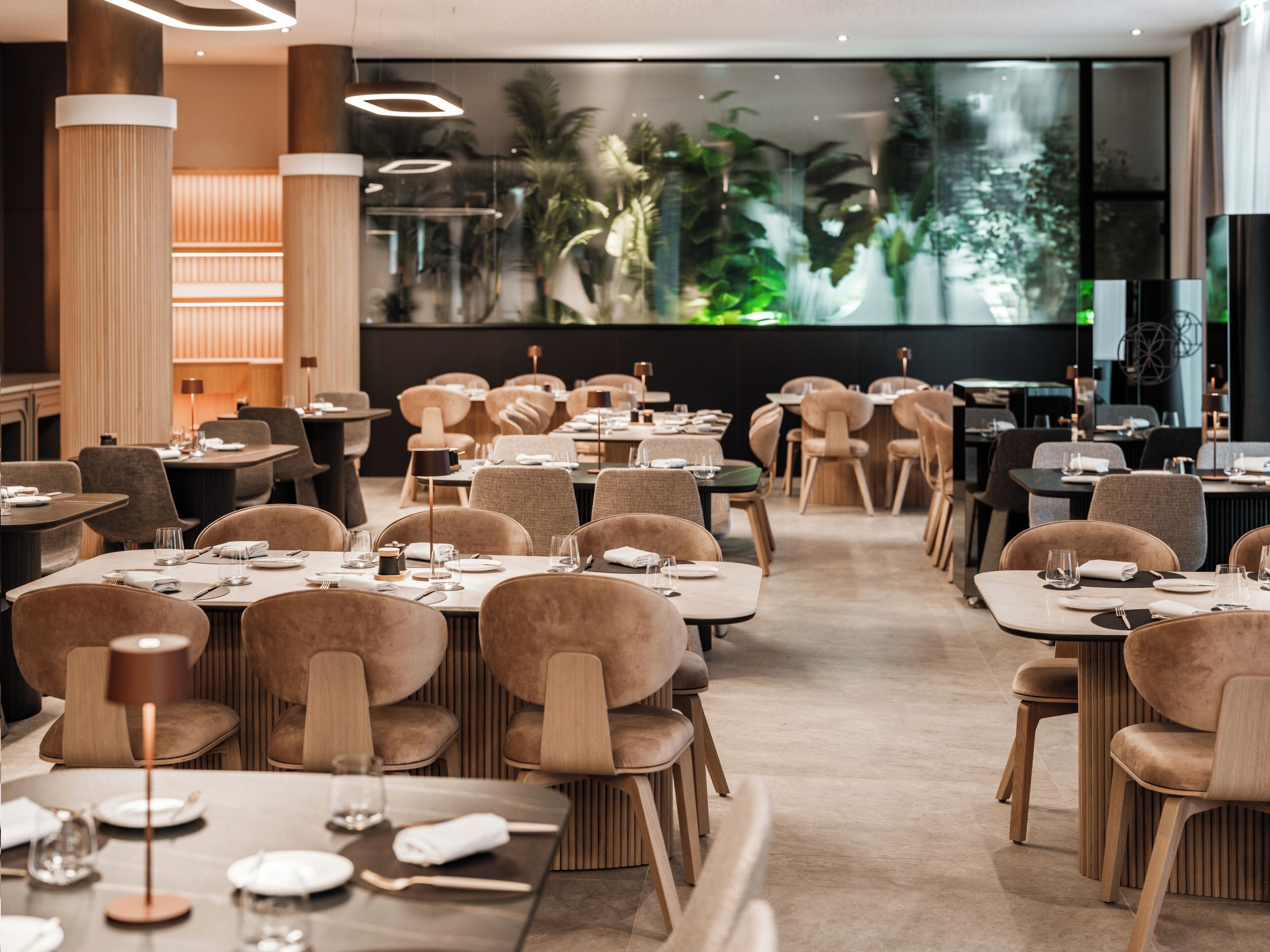 A modern restaurant with multiple dining tables and chairs, large windows with a view of greenery, and contemporary lighting fixtures.