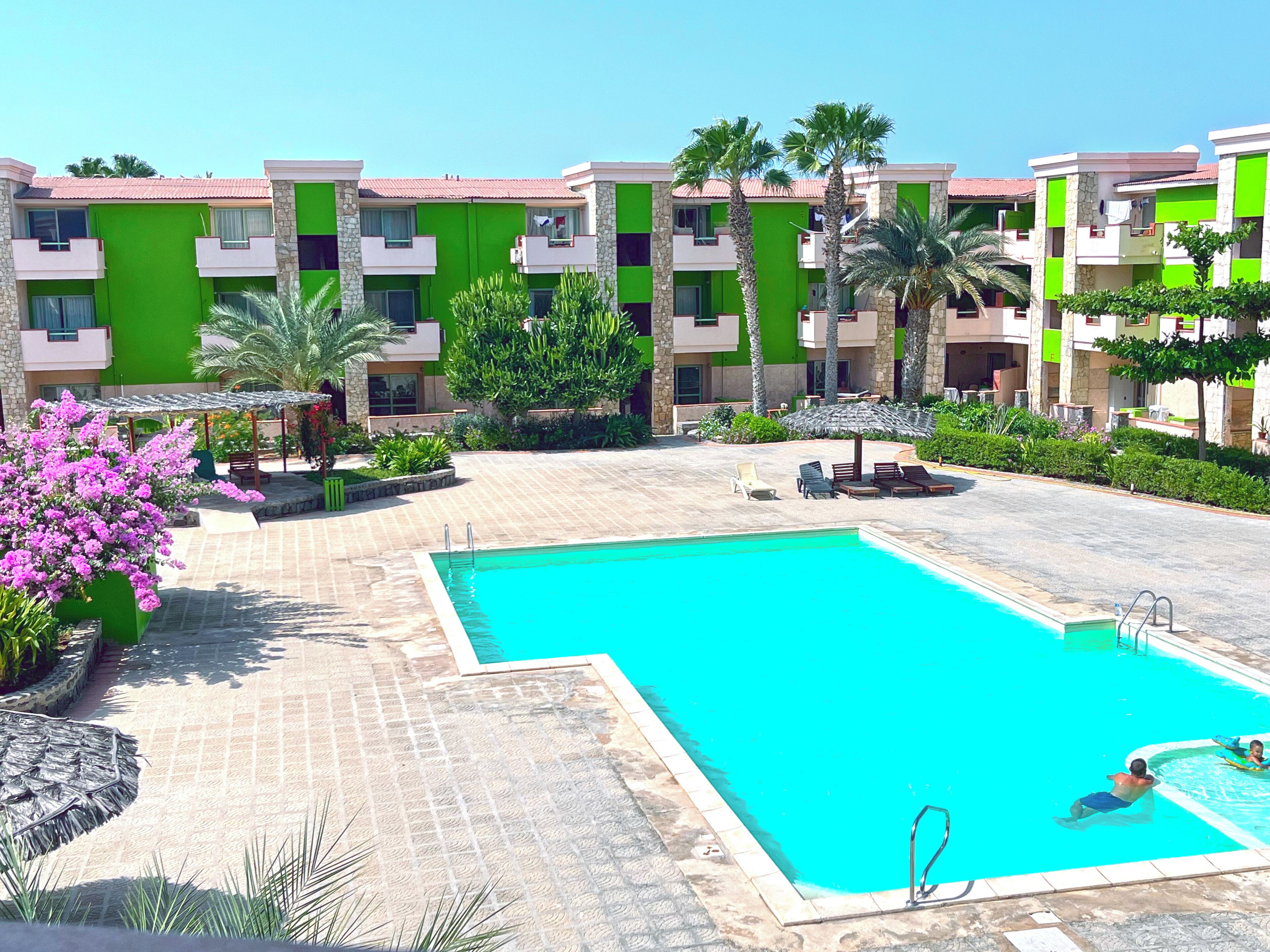 A resort with a swimming pool, palm trees, and a green and beige building facade.