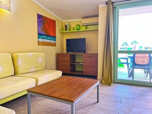 Ein Wohnzimmer mit einer Couch, einem Couchtisch, einem Fernseher auf einer Holzkommode und einer Glastür, die zu einem Außenbereich mit Sitzgelegenheiten führt.