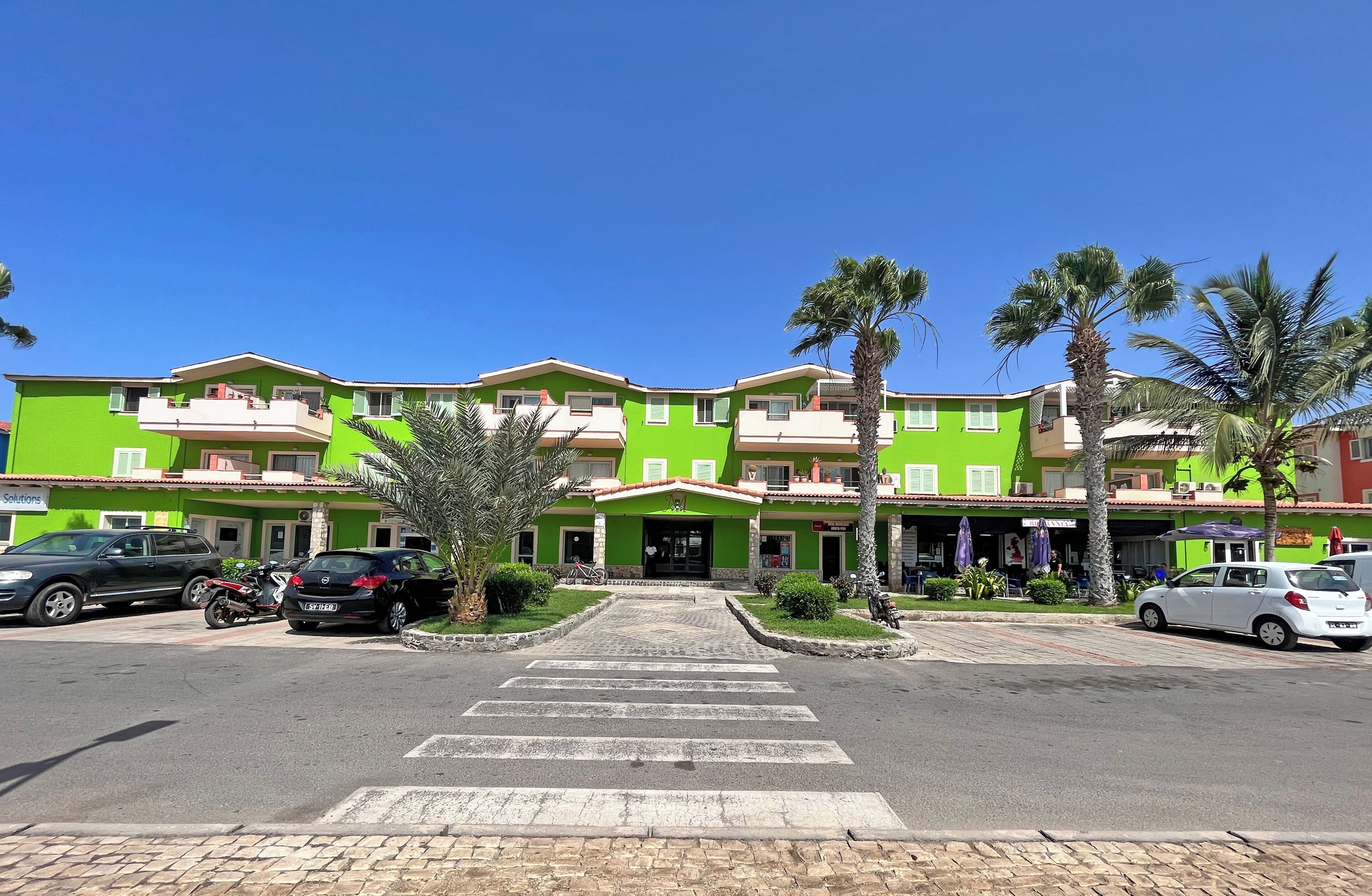 A green multi-story hotel with a central entrance, parking area, and palm trees.