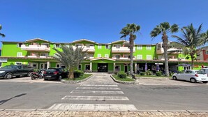 A green multi-story hotel with a central entrance, parking area, and palm trees. - P22B Cozy & Bright, Steps from the Sea (Santa Maria)