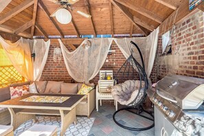 Un espace extérieur aménagé dans un style rustique, avec un canapé en osier, un fauteuil suspendu et un barbecue.