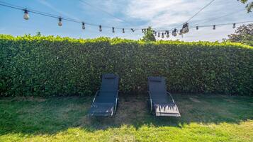 Twee lounge stoelen gericht op een net gesnoeide haag, met lichtjes erboven.