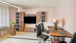 A modern living room with a flat-screen TV, wooden cabinetry, a dining area with a table and chairs, and a potted plant.
