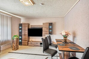 A modern living room with a flat-screen TV, wooden cabinetry, a dining area with a table and chairs, and a potted plant.