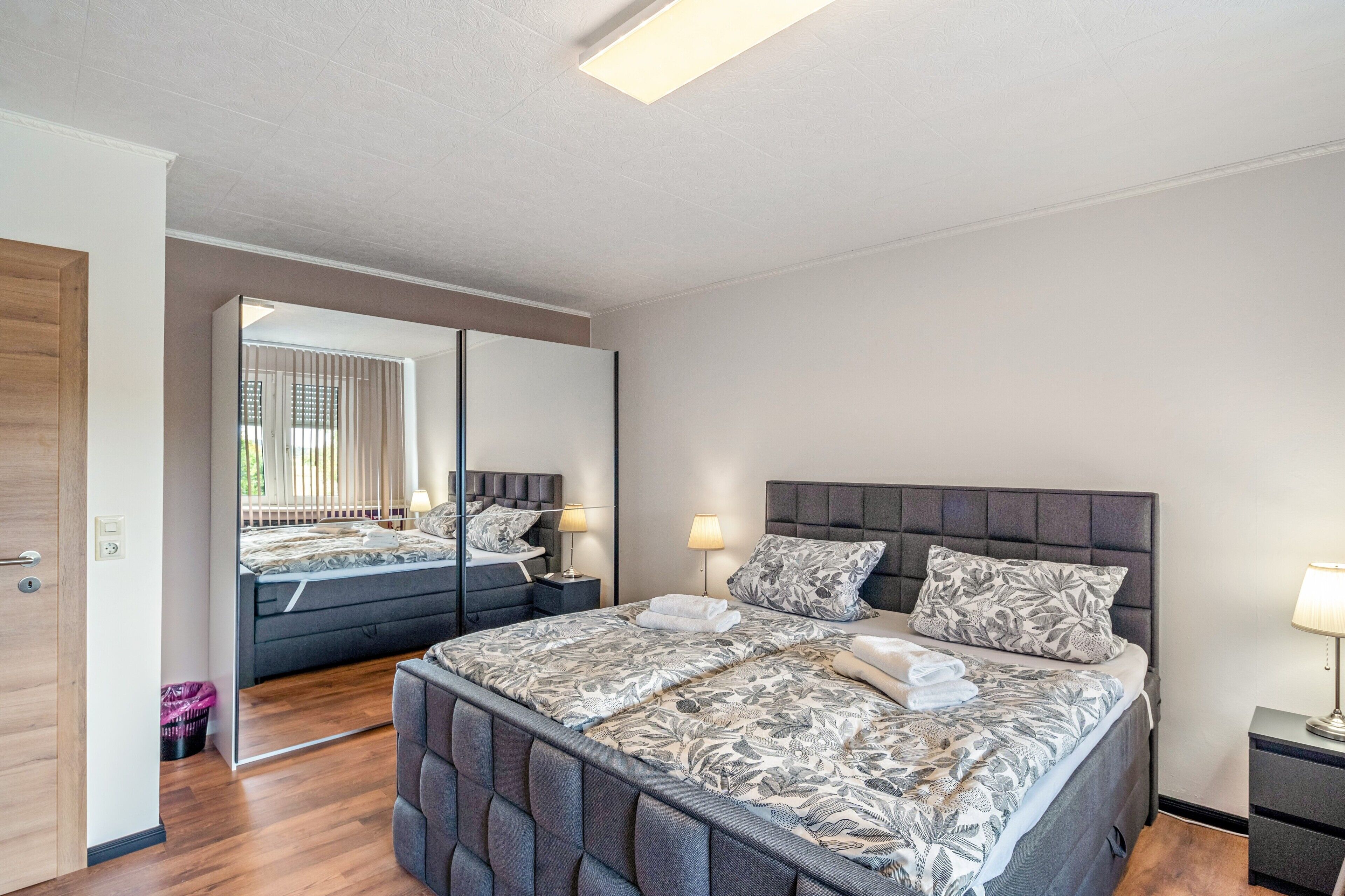 A modern bedroom with a large bed, a nightstand, a mirror, and a wooden door.