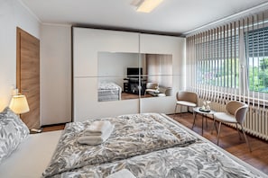 A modern hotel room with a large bed, a seating area with chairs, a small table, and a large window with blinds. - Apartment "Landleben 2" with Shared Terrace, Private Garden, and Wi-Fi (Erkelenz)