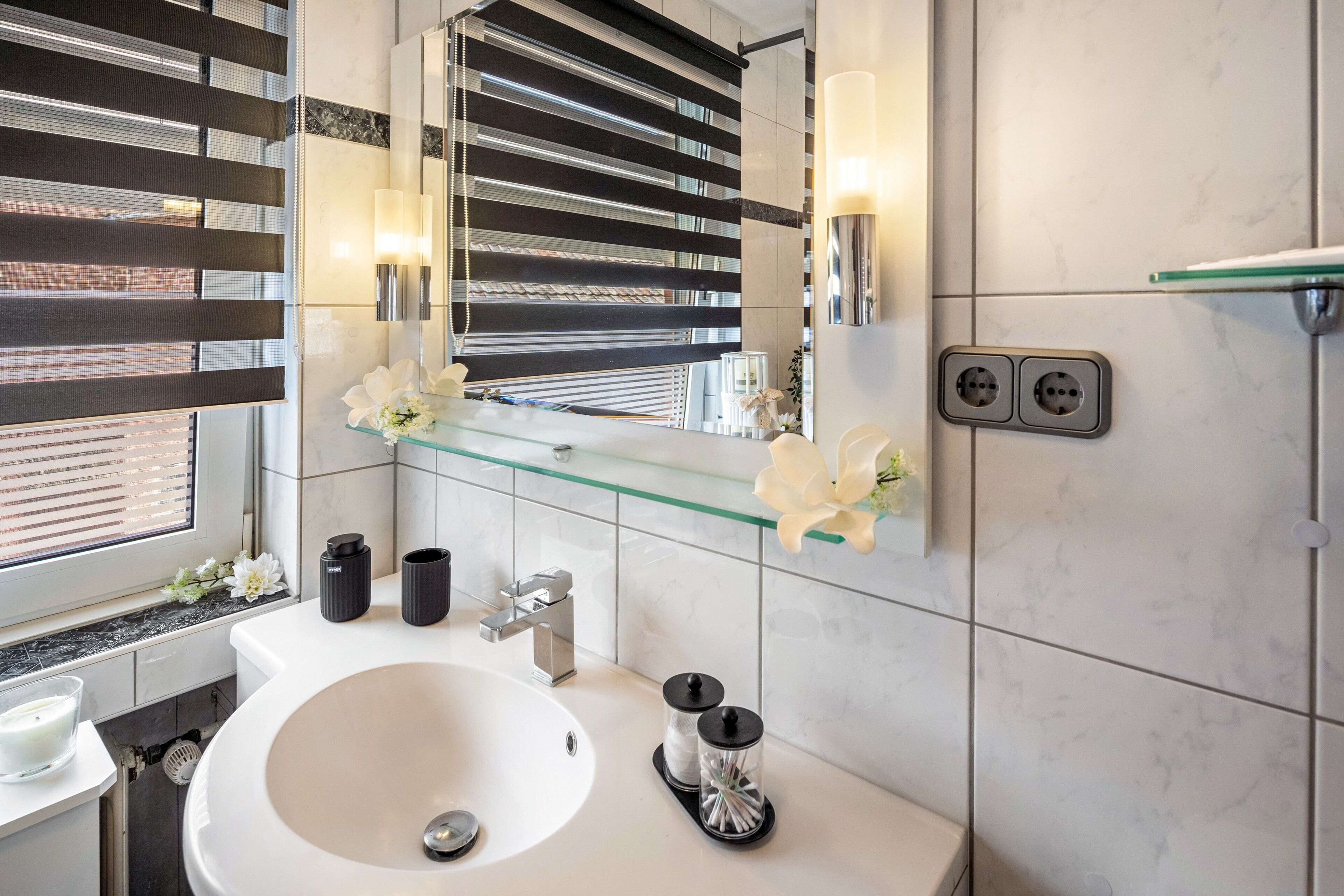 A modern bathroom with a large mirror, a glass shelf with flowers, a sink, and a toilet.
