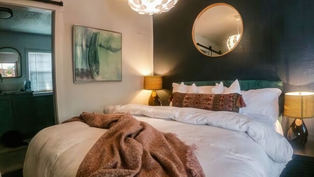 A bedroom with a large bed, a round mirror, a painting, and a unique ceiling light fixture.