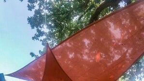 Une maison à l’extérieur sombre, un espace de détente extérieur abrité avec un canapé et des fauteuils blancs, et une grande voile d’ombrage rouge.