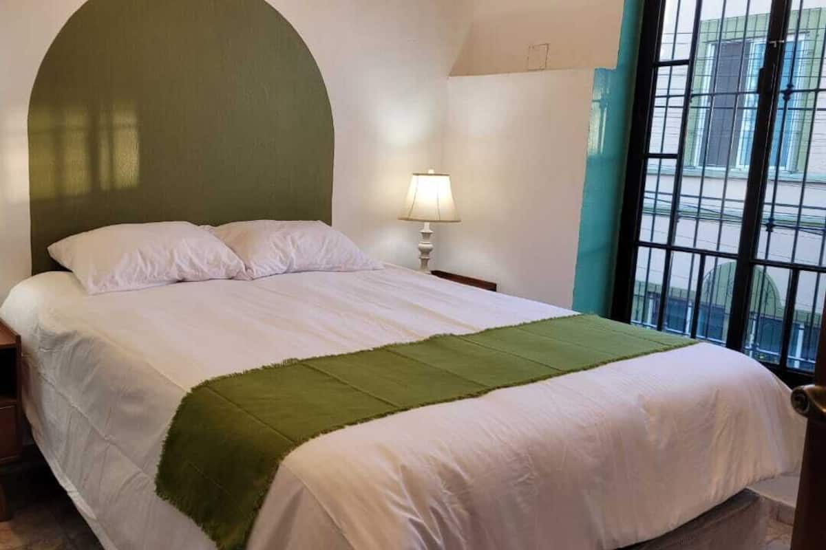 Un llit amb una manta verda, dues làmpades de nit, una tauleta de nit i una finestra amb vistes.
