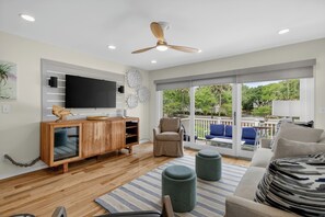 Ein modernes Wohnzimmer mit Flachbildfernseher, Holzschrank, beiger Couchgarnitur, Hocker und Deckenventilator.