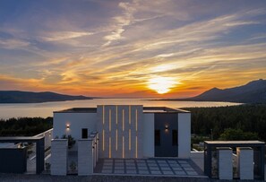 Ein modernes Gebäude mit minimalistischem Design, einem gepflasterten Eingang und Blick auf einen Sonnenuntergang über Wasser und Berge.