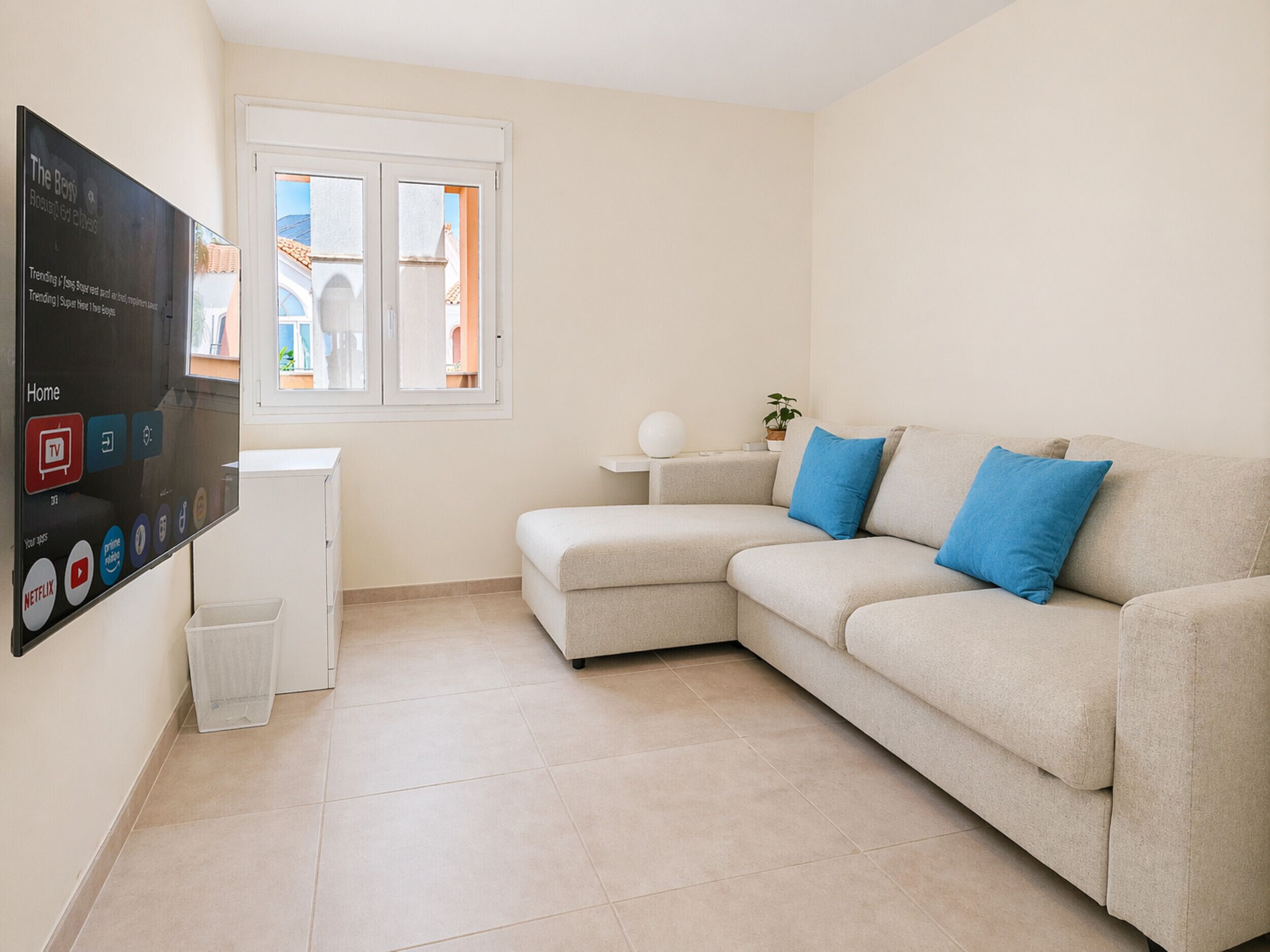 A living room with a beige sofa, a small white table, a TV mounted on the wall, and a window with a view of buildings.