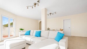 Uma sala de estar moderna com um sofá modular, um pequeno banquinho, uma planta em vaso e um espaço de jantar com mesa posta para dois.