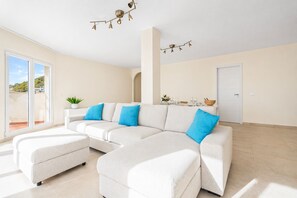 Ein modernes Wohnzimmer mit einer Eckcouch, einem kleinen Hocker, einer Topfpflanze und einem Essbereich mit Tisch für zwei Personen.
