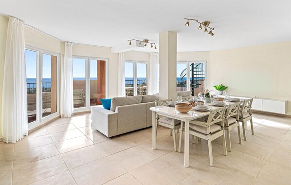 Ein geräumiges Wohnzimmer mit Sofa, Esstisch und Stühlen, großen Fenstern mit Vorhängen und Blick aufs Meer.