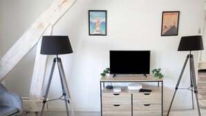 Moderni olohuone, jossa on televisio, kaksi lattiavalaisinta, puinen työpöytä ja kaksi seinälle kiinnitettyä julistetta.