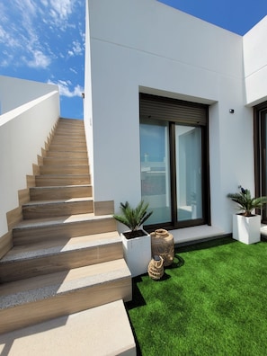 Una casa moderna con una escalera que lleva a un balcón, un gran ventanal corredero y un pequeño césped con plantas en macetas.