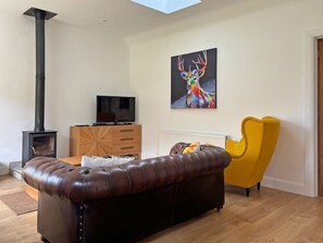 Un salon avec un canapé en cuir marron, un fauteuil jaune, un téléviseur posé sur un meuble en bois, une cheminée et un tableau représentant un cerf.