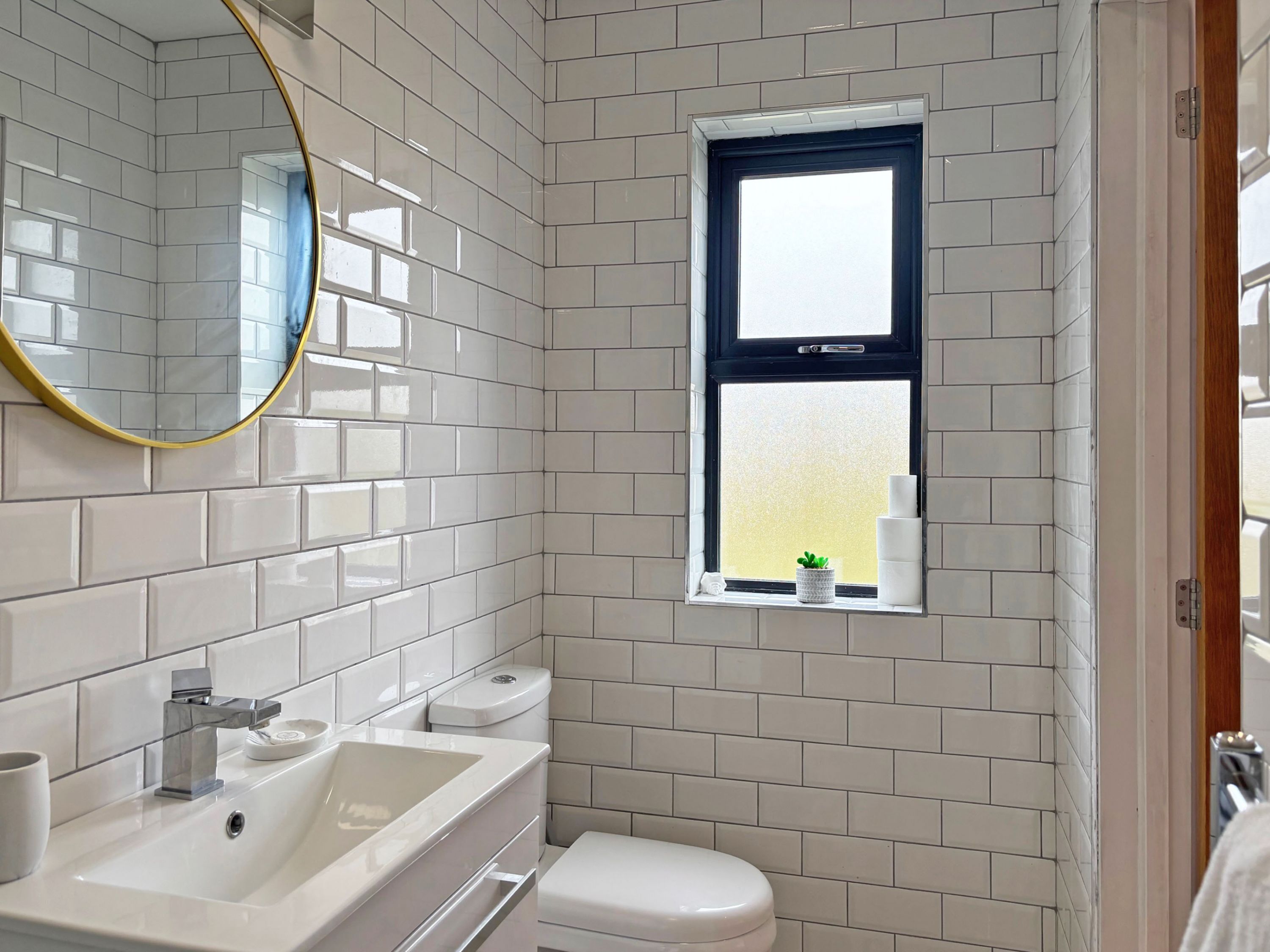 A bathroom with white tiled walls, a window with a view, a sink with a mirror, and a toilet.