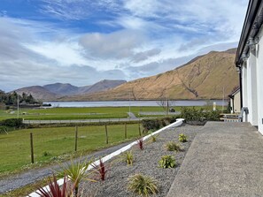 On voit un bâtiment avec une zone pavée devant, un champ d’herbe et des montagnes en arrière-plan.