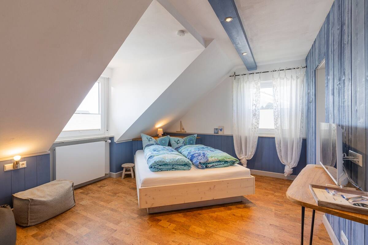A bedroom with a sloped ceiling, a bed with patterned bedding, a small stool, a table with a computer, and a window with curtains.