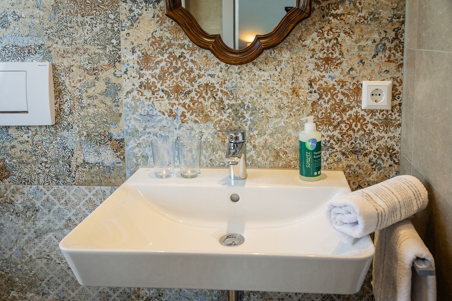 Une salle de bain avec un lavabo blanc, deux verres transparents, un flacon de savon liquide, une serviette et un miroir au mur.