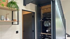Une buanderie moderne dotée d’un lave-linge et d’un sèche-linge encastrés, d’un comptoir en bois et d’un miroir placé au-dessus d’une étagère où sont disposés des objets décoratifs.
