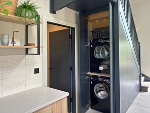 A modern laundry room with a built-in washer and dryer, a wooden counter, and a mirror above a shelf with decorative items. - Cottage 505 - Roundstone - sleeps 8 guests  in 4 bedrooms (Roundstone)