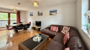 A living room with a sofa, a coffee table, a TV, and a dining area with chairs and a table. - Apartment 514 - Clifden - sleeps 6 guests  in 3 bedrooms (Clifden)