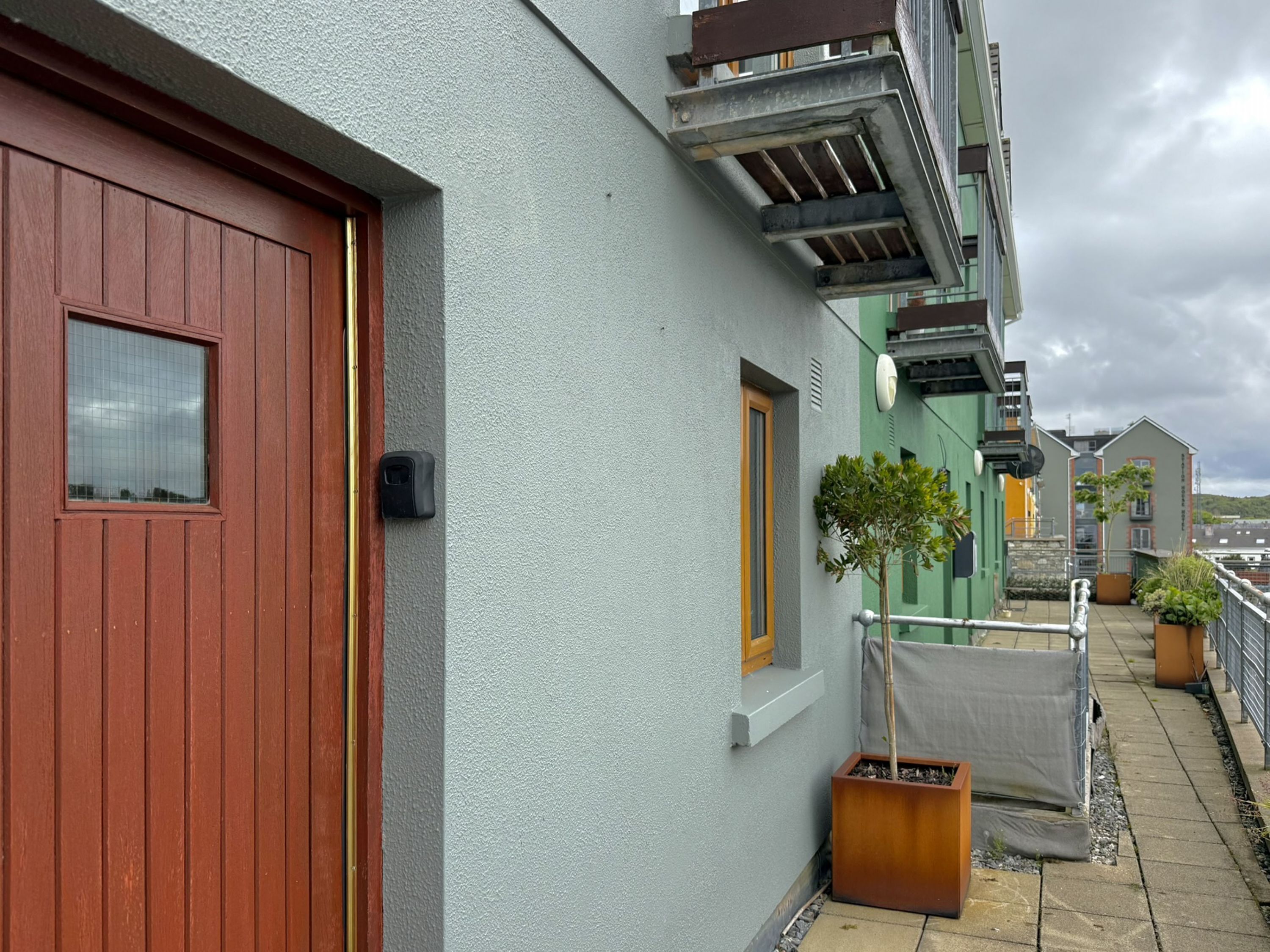 Een woongebouw met een rode deur, een klein raam, een balkon met een bloempot en een grijze buitenmuur.