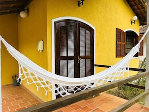 A yellow building with a wooden door and window shutters, a hammock, and a brick floor.