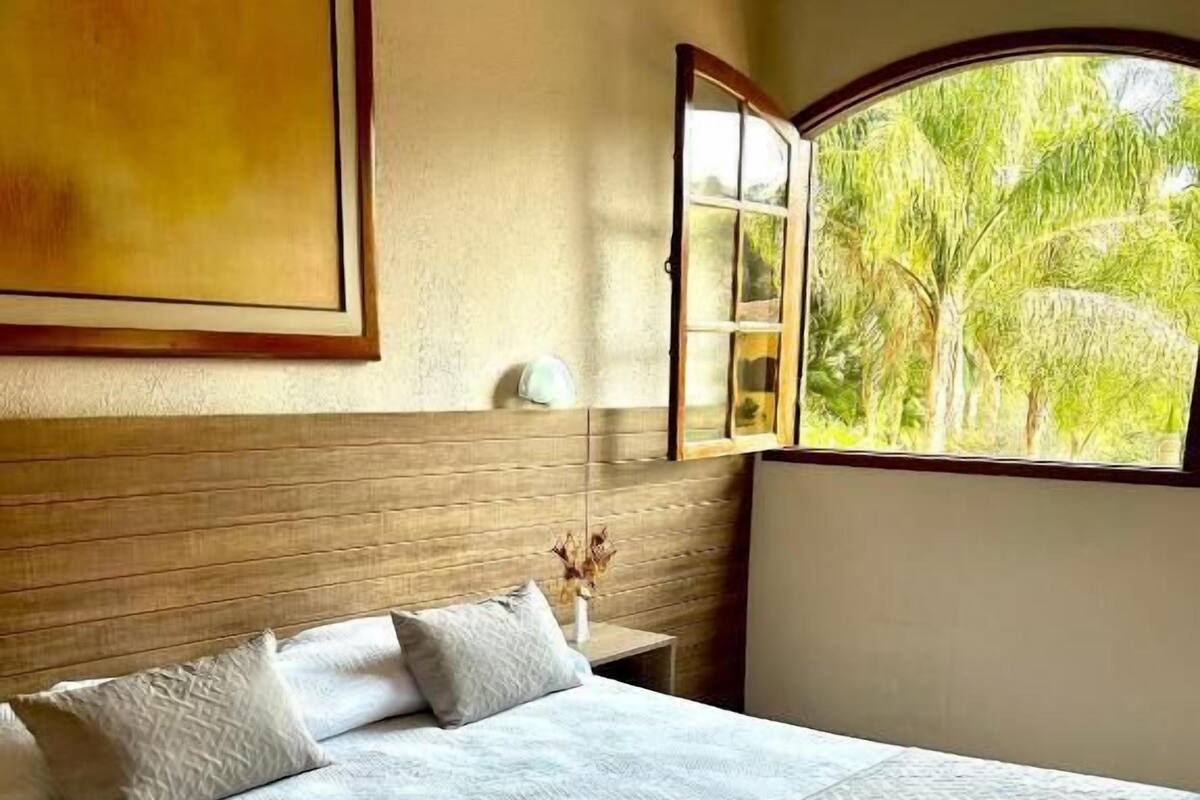 Un dormitorio con una cama grande, una mesita de noche con lámpara, una ventana que da a vistas de vegetación y un cuadro en la pared.