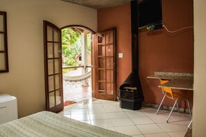 Una habitación con cama, una mesita, televisor, chimenea y vista a un jardín desde una puerta abierta.