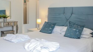 Una cama bien hecha con sábanas blancas y dos almohadas azules, una mesita de noche con una lámpara y un espejo en la pared.