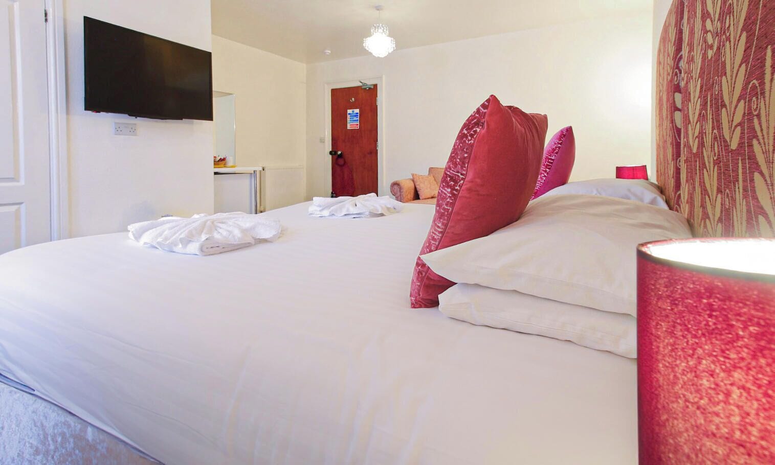 A hotel room with a bed, a flat-screen TV mounted on the wall, a door, and a bedside lamp.