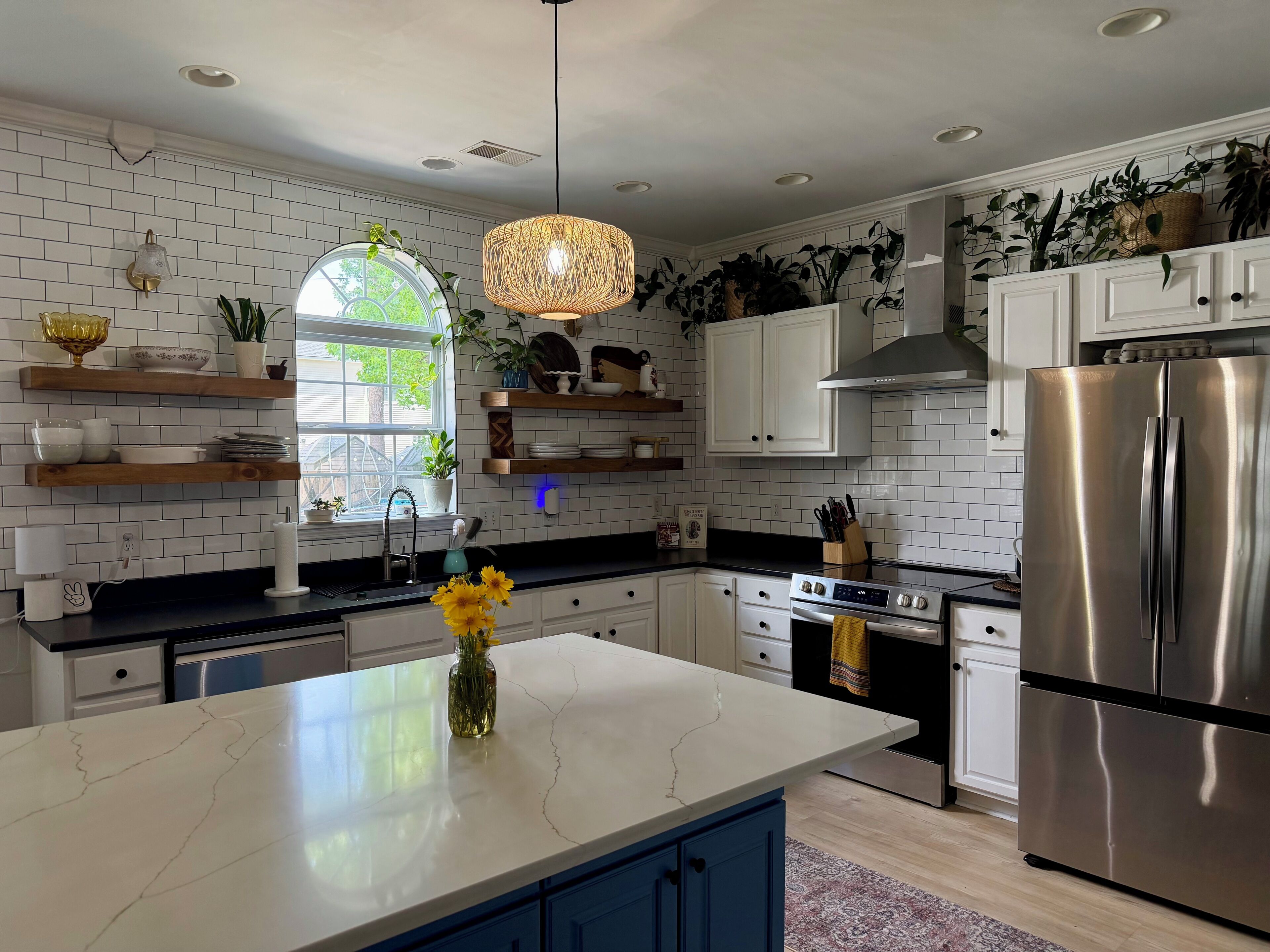 Una cocina moderna con azulejos blancos tipo metro, una isla central, electrodomésticos de acero inoxidable y plantas decorativas.