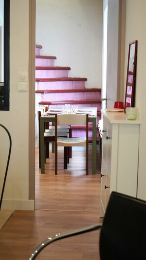Ein Essbereich mit einem Holztisch und Stühlen, eine Treppe mit rosafarbenen Stufen sowie ein weißes Schränkchen mit Spiegel.