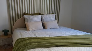 Ein Bett mit gestreifter Kopfteil, zwei Kissen mit Muster, eine grüne Tagesdecke und ein kleiner runder Tisch mit einem grünen Gegenstand darauf.