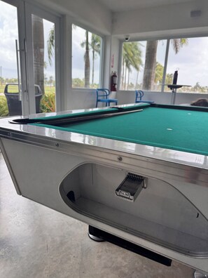 A pool table with a price tag, a trash can, and a view of palm trees through large windows.