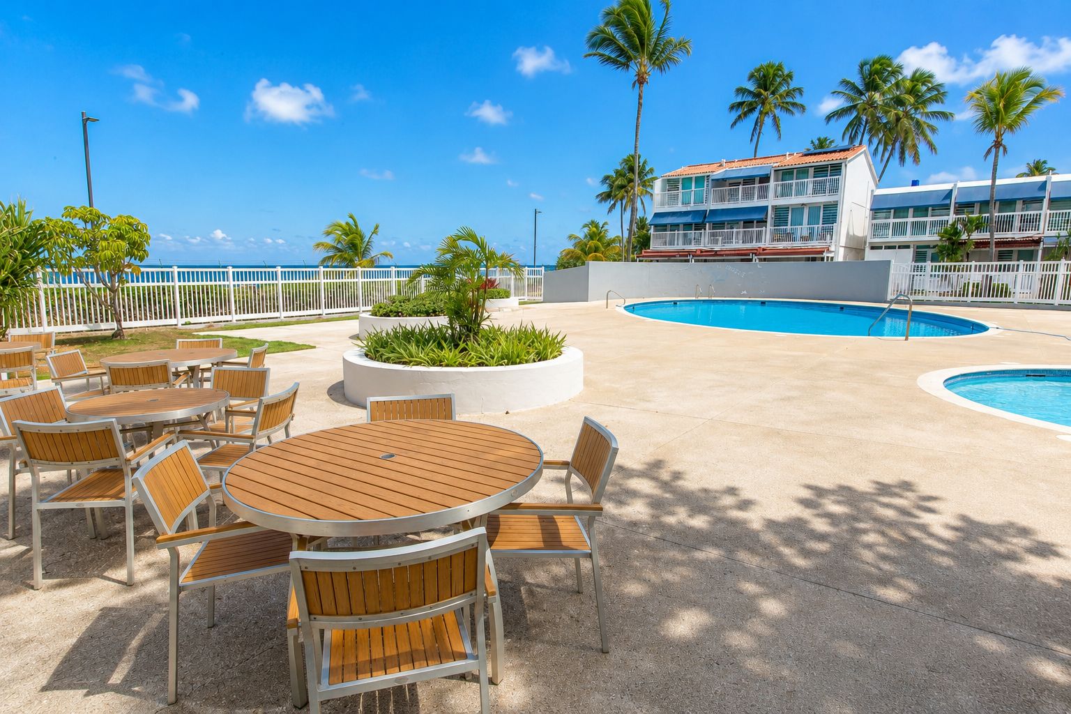 A resort pool area with multiple tables and chairs, a swimming pool, and a building with balconies.