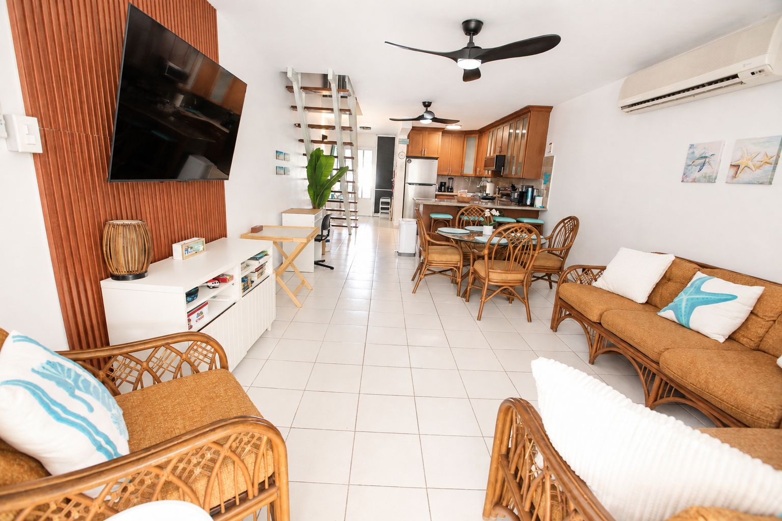 A modern living room with a flat-screen TV, wicker furniture, a dining area with chairs, and a kitchen in the background.