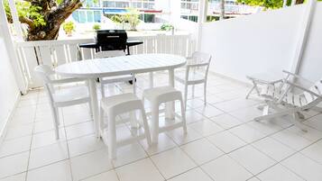 A white outdoor patio with a round table and chairs, a grill, and a view of buildings and trees.