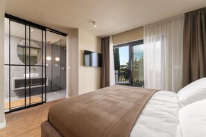 Ein Schlafzimmer mit Bett, Fernseher, einer Schiebetür zum Balkon und einem Badezimmer mit Waschbecken und Spiegel.