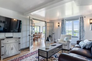A spacious living area with a flat-screen TV, a dining table, a sofa, and a view of the outdoors. - Riverview Countryside Farmhouse, King bed Spa Bath (Paroisse de Saint-Louis)