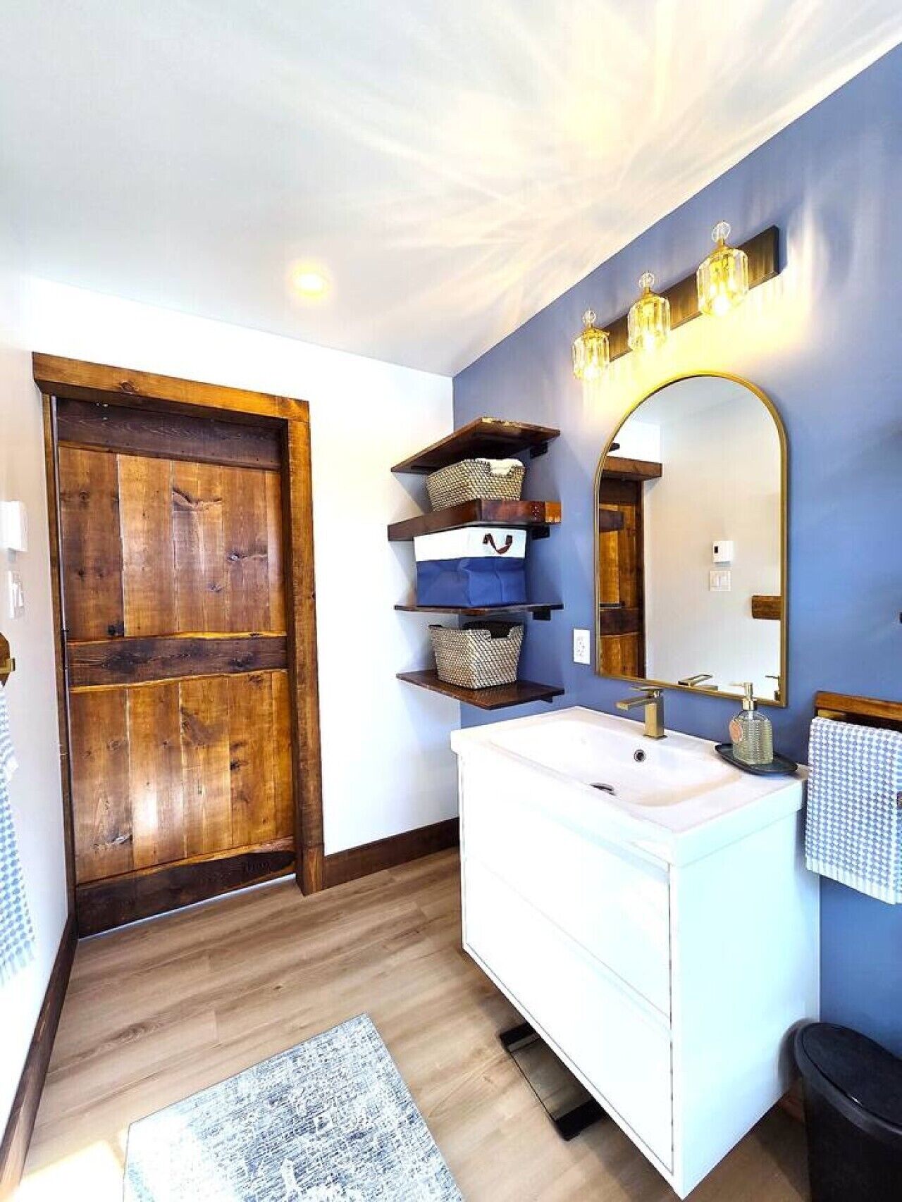 A bathroom with a wooden door, white sink, shelves with baskets, a mirror, and a rug.
