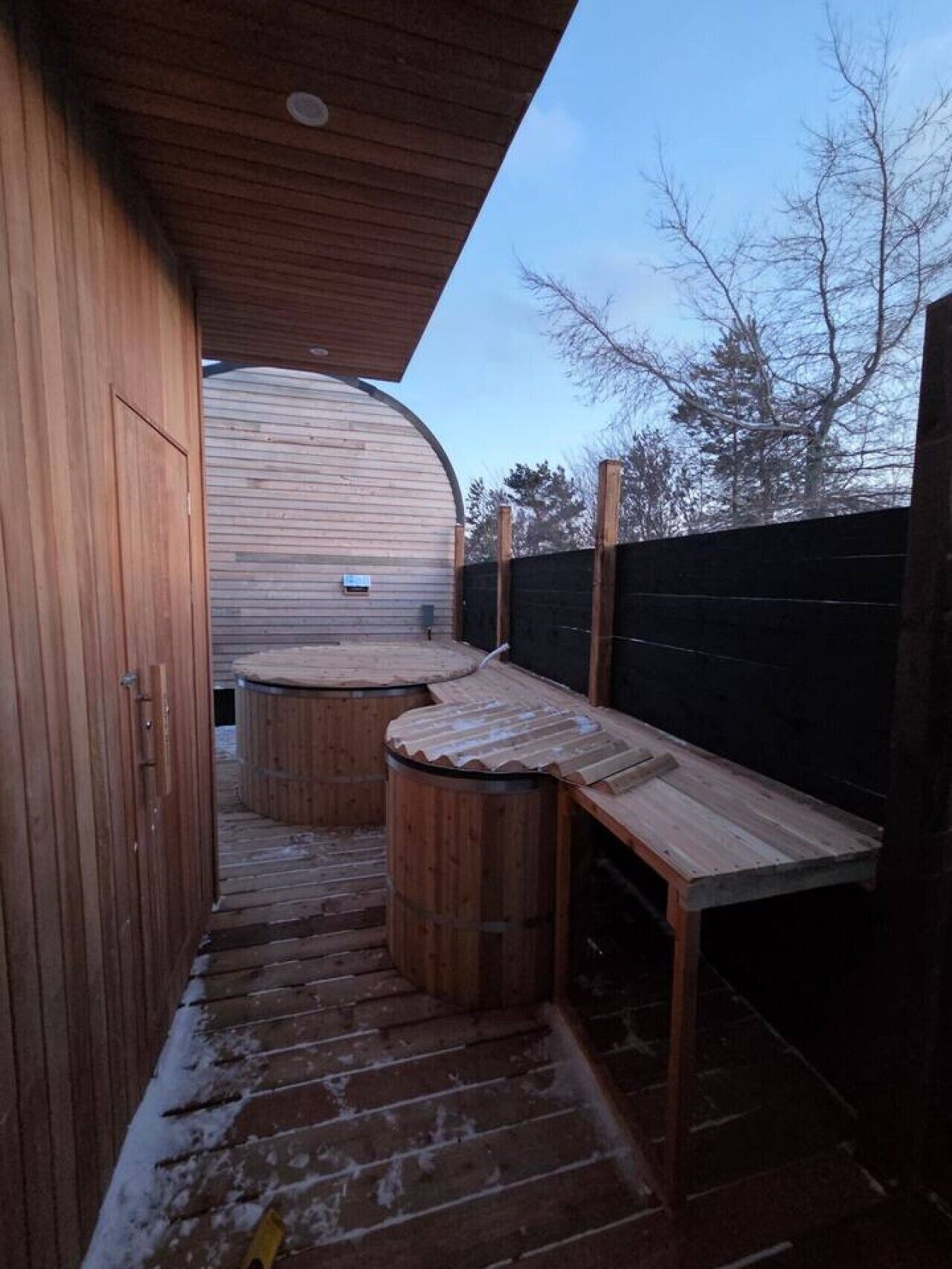 Ett utomhusområde med trägolv, en möblerad sittplats under tak, ett träbord och en tunna.